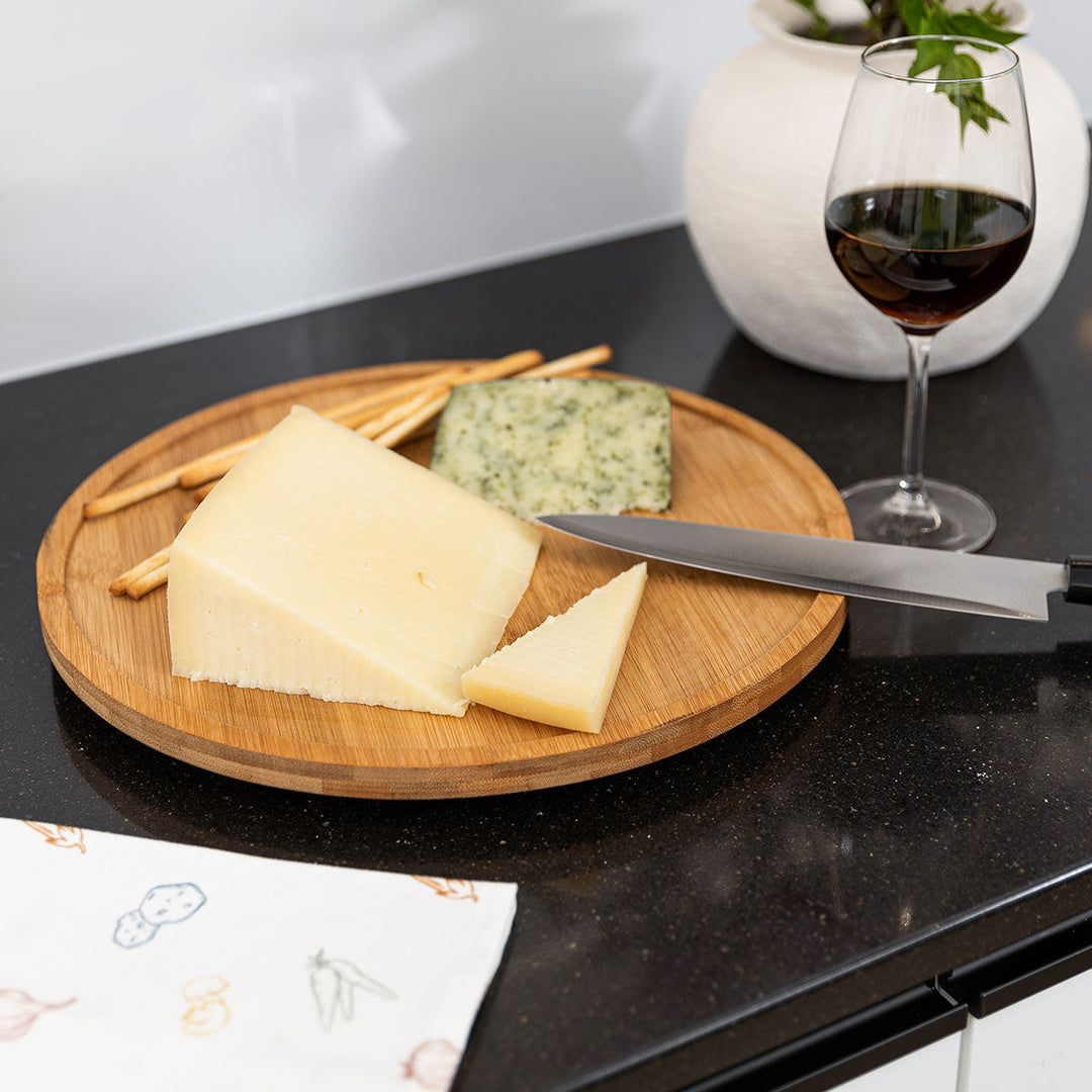 Ξύλινη στρογγυλή βάση Bamboo Essentials Estia σε χρήση με ποτήρι κρασί και διάφορα τυριά σε μαύρο πάγκο κουζίνας.