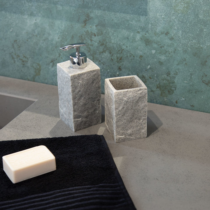 Γκρι stone dispenser με ασημί αντλία σε μοντέρνο μπάνιο, δίπλα σε γκρι ποτήρι και σκούρα πετσέτα πάνω σε πάγκο.