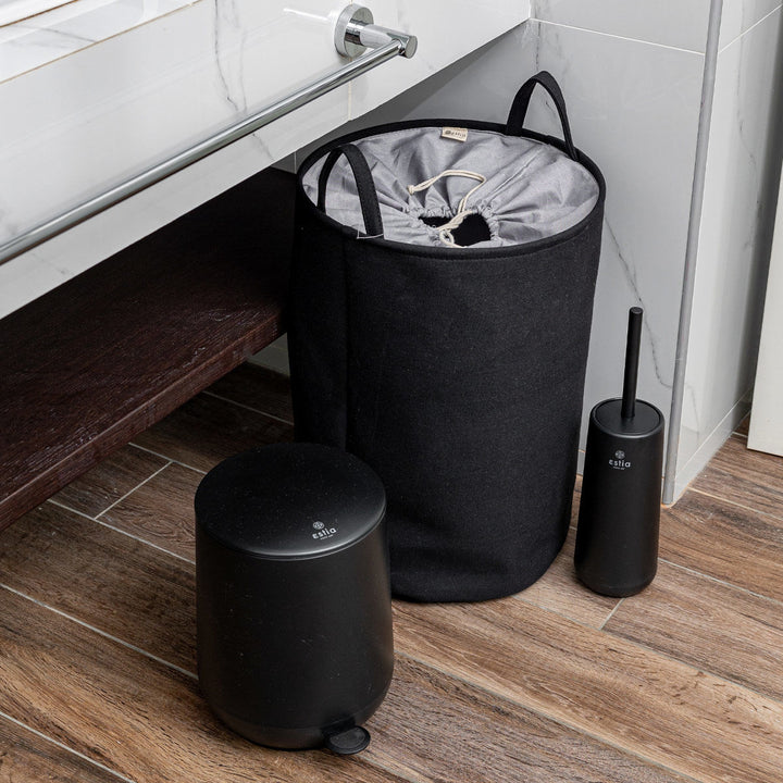 Μαύρος κάδος απορριμμάτων, υφασμάτινο καλάθι και πιγκάλ μπάνιου σε ξύλινο δάπεδο, δίπλα σε λευκό νιπτήρα και μαρμάρινα πλακάκια.
