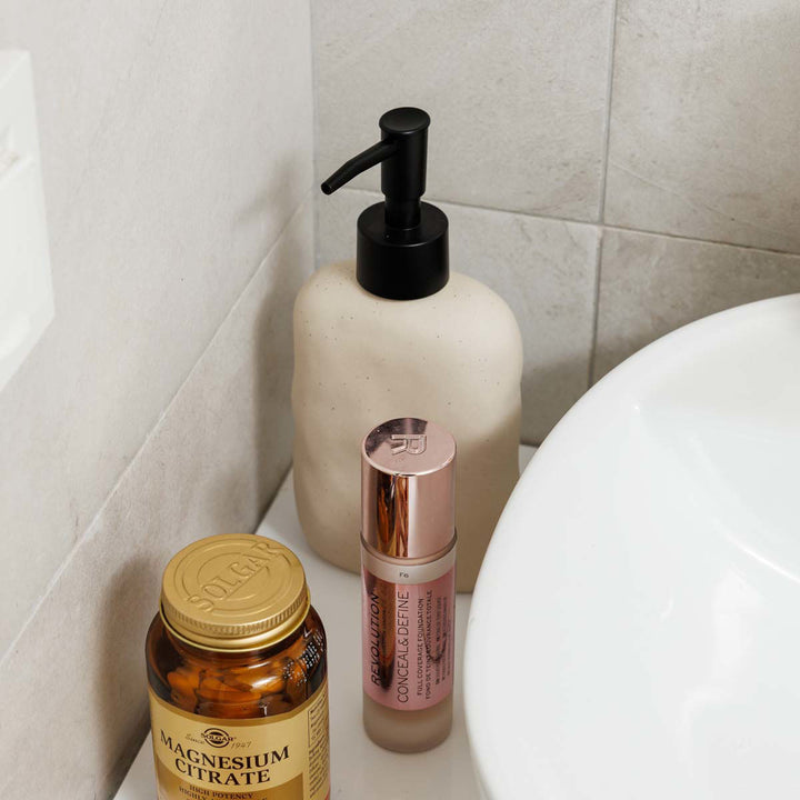 Dispenser σαπουνιού σε ανοιχτό μπεζ χρώμα με μαύρη αντλία, τοποθετημένος σε μπάνιο δίπλα σε καλλυντικά και λευκό νιπτήρα.