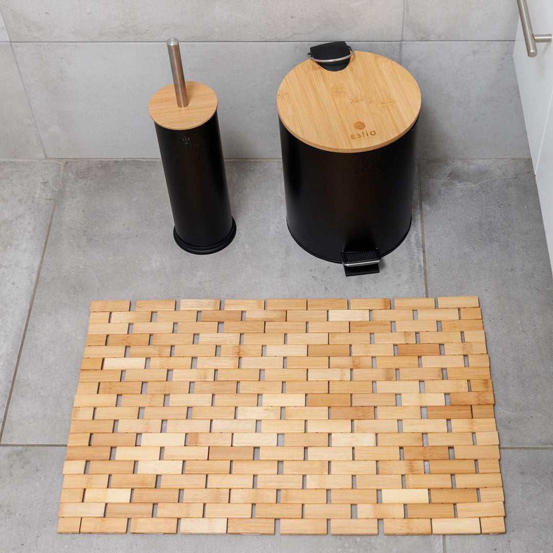 Ξύλινο πατάκι από bamboo τοποθετημένο σε μπάνιο με γκρι πλακάκια, δίπλα σε μαύρα αξεσουάρ με καπάκι bamboo.