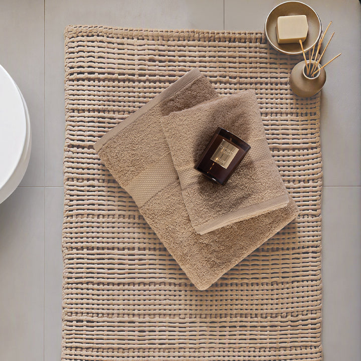 Μπεζ χαλάκι μπάνιου Delight Nef-Nef Homeware τοποθετημένο σε μπάνιο με διπλωμένες πετσέτες και διακοσμητικά αντικείμενα.