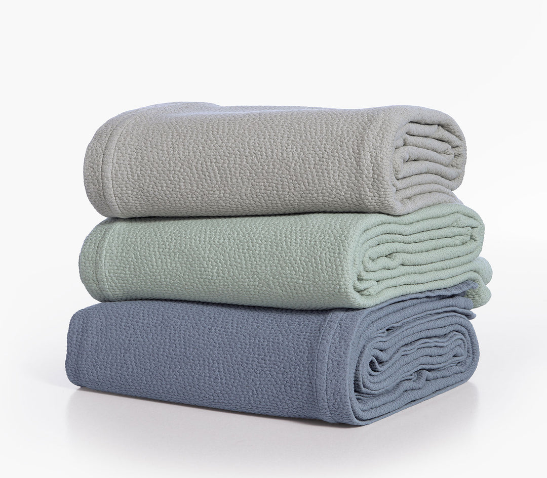 Τρεις διπλωμένες βαμβακερές κουβέρτες Nef-Nef Homeware σε πράσινο, μπεζ και μπλε χρώμα στοιβαγμένες.