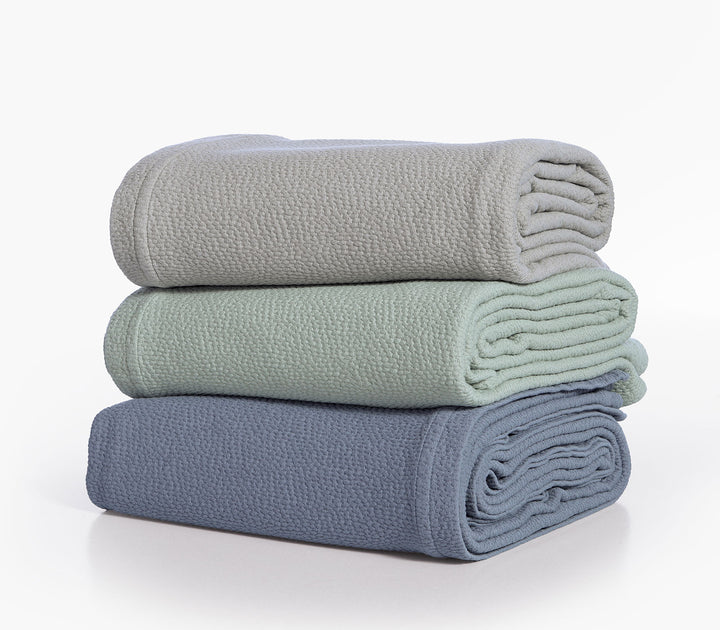 Τρεις διπλωμένες βαμβακερές κουβέρτες Nef-Nef Homeware σε πράσινο, μπεζ και μπλε χρώμα στοιβαγμένες.