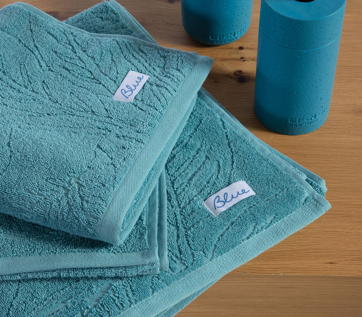 Πετρόλ βαμβακερές πετσέτες Nef-Nef Homeware με ανάγλυφο φυλλωτό σχέδιο τοποθετημένες σε ξύλινη επιφάνεια δίπλα σε μπλε αξεσουάρ μπάνιου.