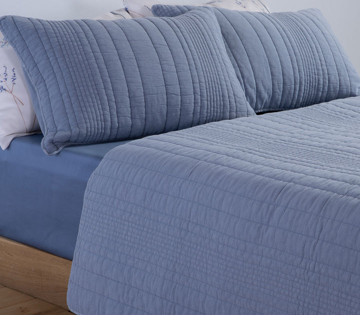 Μπλε κουβερλί Nef-Nef Homeware σε κρεβάτι με ασορτί μαξιλάρια και λευκά σεντόνια με διακριτικό φλοράλ σχέδιο.