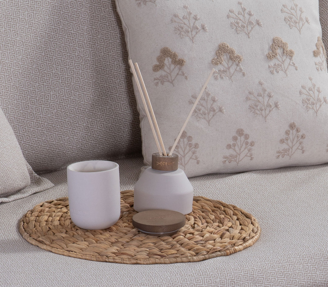 Αρωματικό χώρου με στικς Cotton Flower της Nef-Nef Homeware δίπλα σε αρωματικό κερί πάνω σε ψάθινο σουπλά σε καναπέ.
