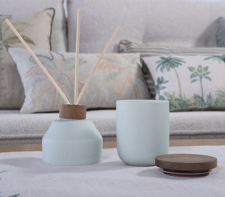 Αρωματικό χώρου με στικς Gardenia της Nef-Nef Homeware πάνω σε τραπέζι σαλονιού μπροστά από καναπέ με διακοσμητικά μαξιλάρια.