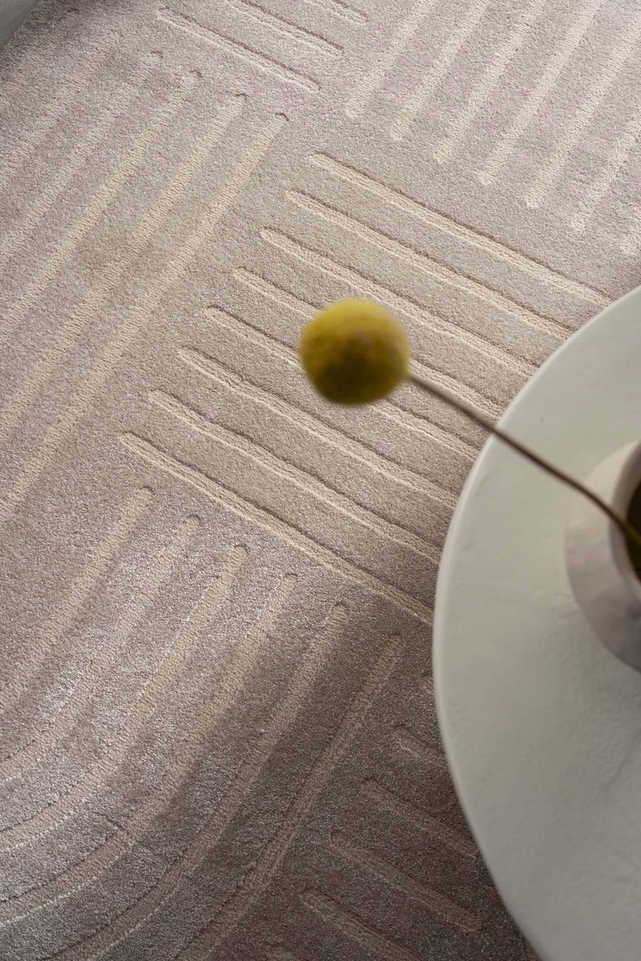Μπεζ χαλί Organic Royal Carpet με καμπυλωτές γραμμές και ανάγλυφο σχέδιο, λεπτομέρεια διακόσμησης με βάζο και λουλούδι.