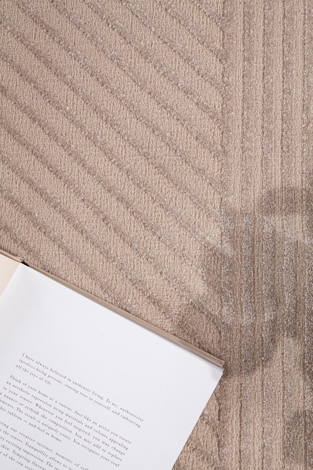 Μπεζ χαλί Organic Royal Carpet με μοτίβο από διαγώνιες και κάθετες γραμμές, με βιβλίο τοποθετημένο επάνω στο πάτωμα.