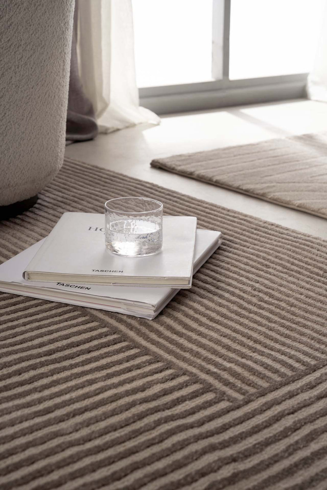Μοντέρνο χαλί Organic Royal Carpet σε μπεζ χρώμα με γεωμετρικό σχέδιο, κοντινή λεπτομέρεια με βιβλία και γυάλινο ποτήρι στο πάτωμα.