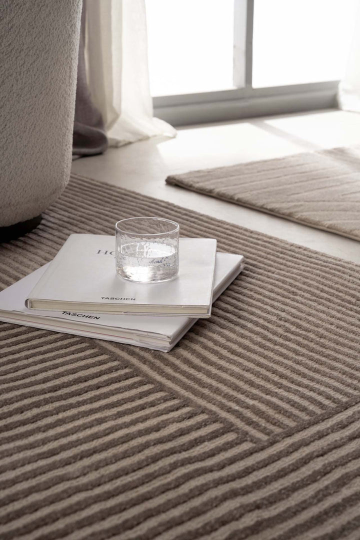 Μοντέρνο χαλί Organic Royal Carpet σε μπεζ χρώμα με γεωμετρικό σχέδιο, κοντινή λεπτομέρεια με βιβλία και γυάλινο ποτήρι στο πάτωμα.