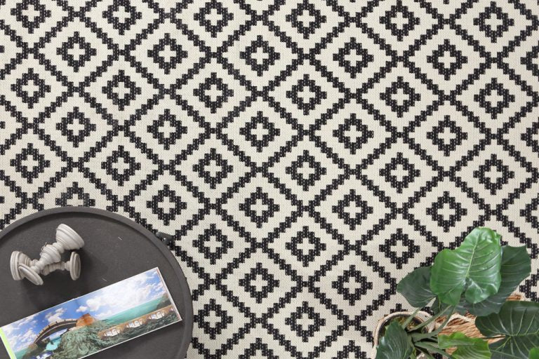 Κοντινή προβολή σε ψάθινο χαλί Flox Royal Carpet με γεωμετρικό μοτίβο δίπλα σε διακοσμητικά και πράσινο φυτό.
