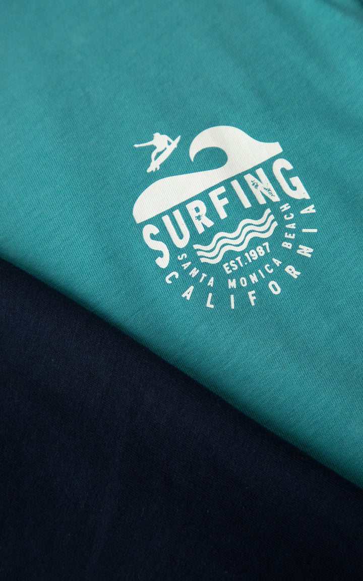Κοντινή λήψη παιδικής πιτζάμας Minerva με surfing σχέδιο σε μπλε βαμβακερή μπλούζα και μαύρη βερμούδα.
