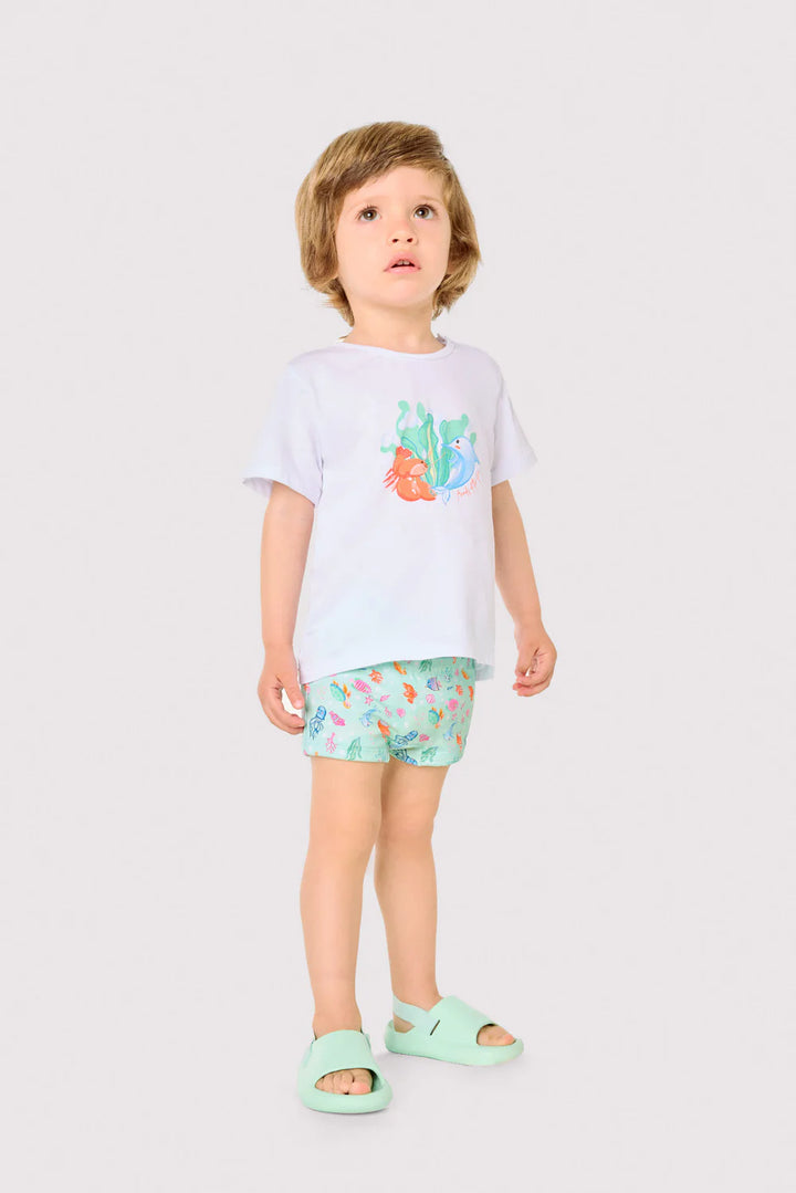 Παιδί φοράει σετ παραλίας με λευκό t-shirt και τιρκουάζ μαγιό με θαλάσσια σχέδια σε ουδέτερο φόντο.