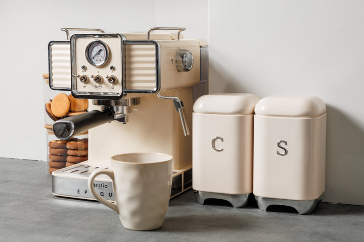 Vintage μηχανή espresso σε εκρού απόχρωση πάνω σε πάγκο κουζίνας, με ποτήρι μπισκότων και δοχεία καφέ, ζάχαρης.